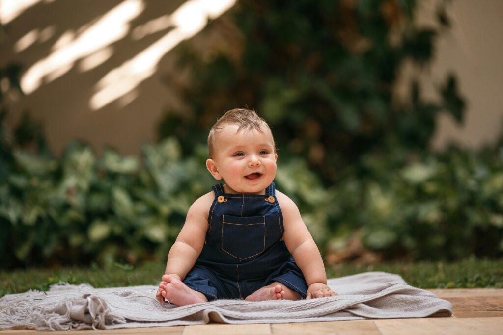 Cute baby in denim sitting on a blanket, smiling in a sunny outdoor setting.