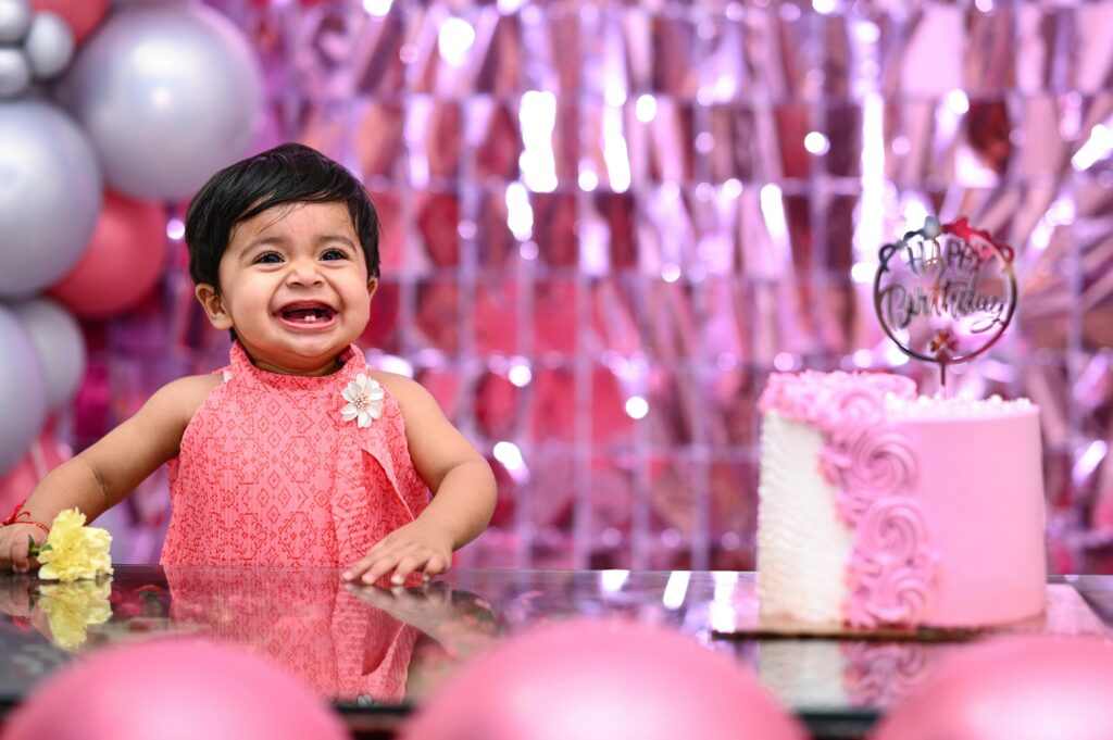 Adorable baby smiling with a cake and festive pink decorations at her first birthday celebration.