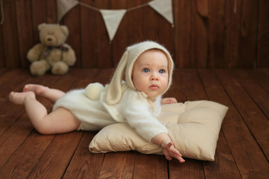 Cute baby in a bunny outfit lying on a soft pillow with a teddy bear in the background.