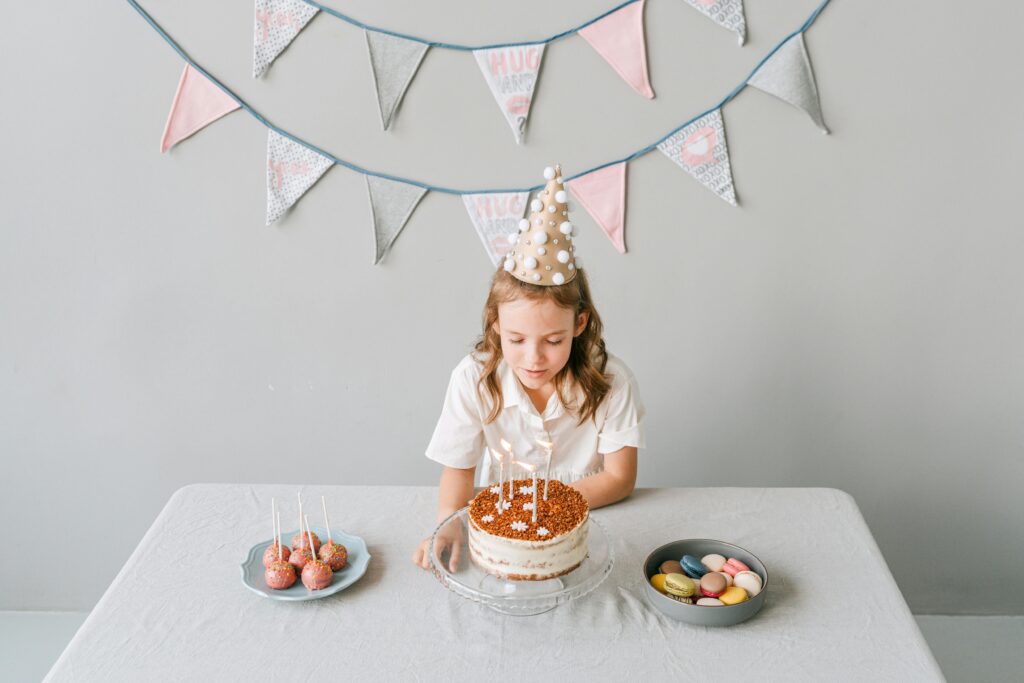 A girl in a party hat making a wish on her birthday cake with macarons on the side.